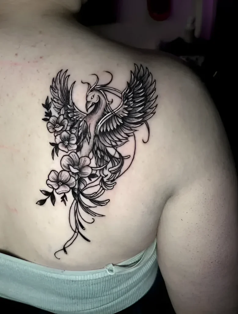 Black and white tattoo of a phoenix with sakura flowers on a girl's shoulder blade, done in a graphic style.