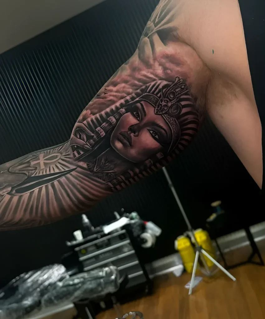 Tatouage réaliste noir et gris de Cléopâtre sur le biceps d'un homme, avec le profil de la reine en coiffe et le symbole Ankh.