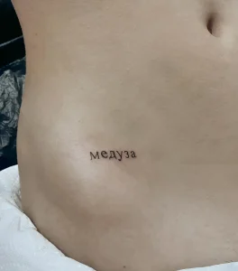 Маленькая татуировка надписью 'медуза' черным цветом на боку девушки, выполненная в минималистичном стиле.