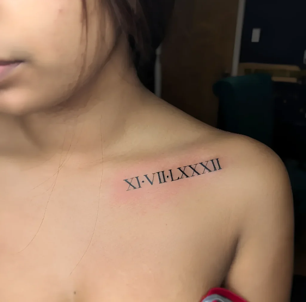 Photo of a collarbone tattoo: black Roman numerals XI-VII-LXXXII, done in a clear line, placed along the collarbone.