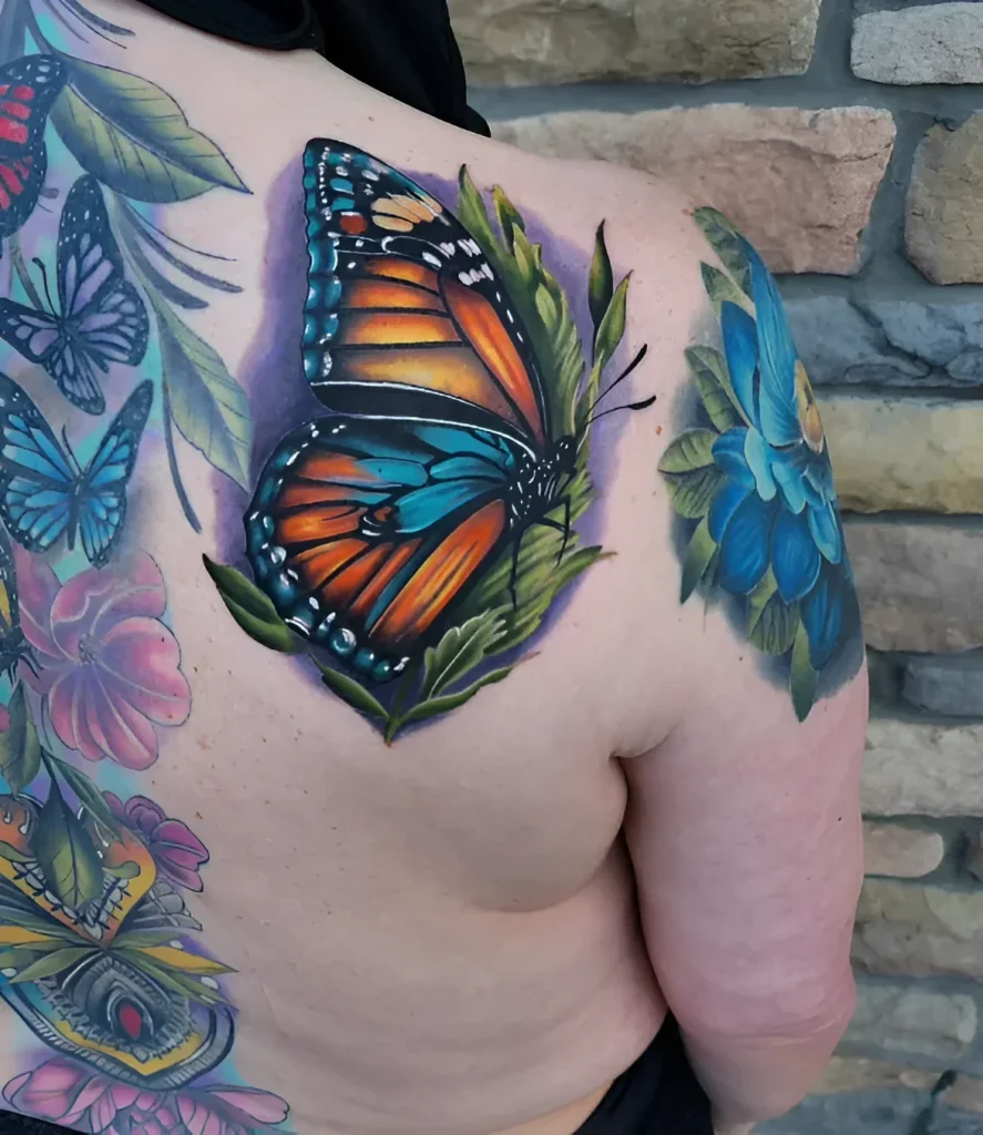 Photo of a tattoo on the shoulder blade: a realistic monarch butterfly with bright orange and black wings, surrounded by green leaves and blue flowers.
