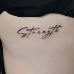 Фотография татуировки надписи 'Strength' каллиграфическим шрифтом на боковой части грудной клетки женщины.