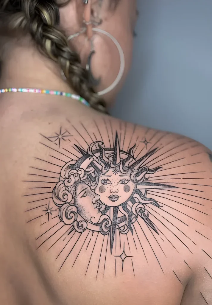 Black and white tattoo on the shoulder blade: a two-faced sun surrounded by rays and decorative elements.