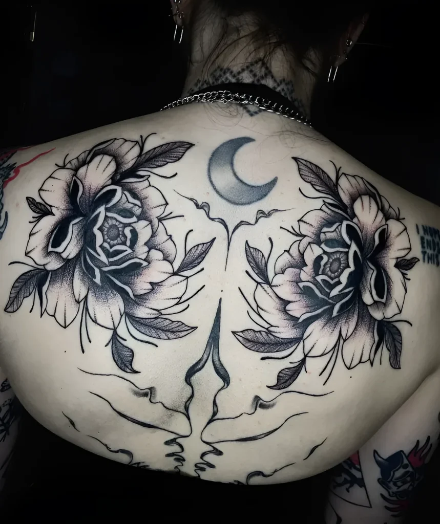 Black and white tattoo on the shoulder blade: two blooming peonies, a crescent moon, and a skull outline, done in a graphic style.
