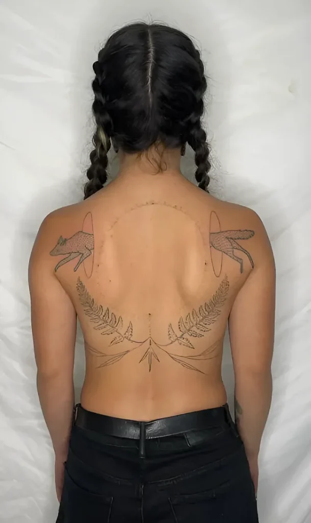 Photo of a tattoo on the shoulder blade: two foxes framed by fern and a moon circle, done in fine graphic technique.