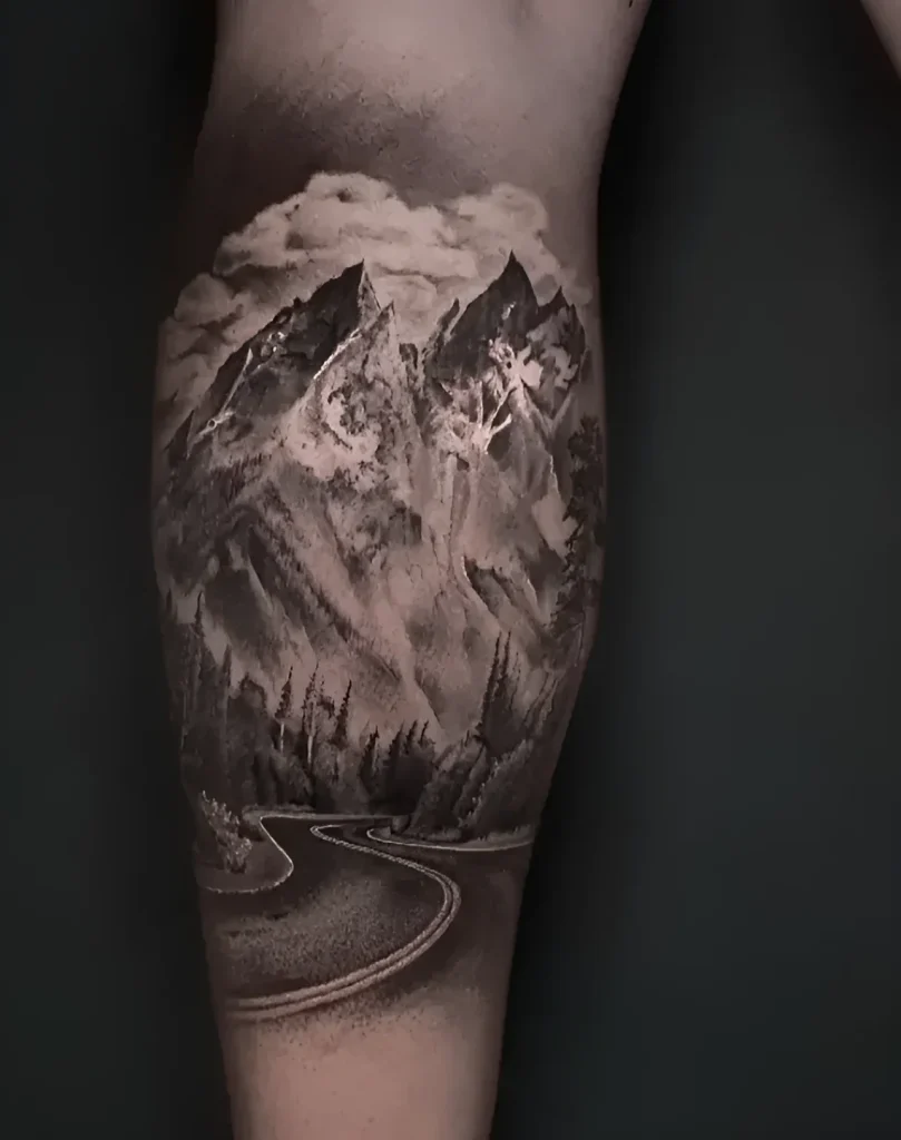 Black and white calf tattoo: majestic mountains covered in clouds, and a winding road stretching into the distance.