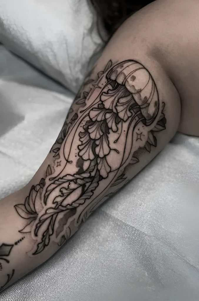 Tatouage noir et blanc d'une méduse sur le biceps, réalisé dans un style graphique avec des éléments végétaux et des étoiles ajoutés.