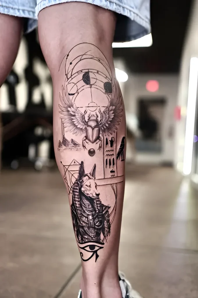 Black and white calf tattoo depicting Anubis, a scarab beetle, and pyramids, done in a graphic style with geometric elements.