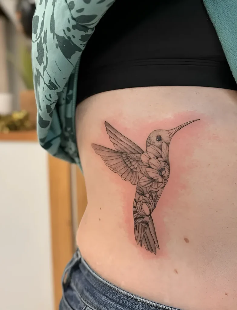 Photographie d'un tatouage de colibri sur le flanc d'une jeune femme, réalisé en technique graphique avec des lignes fines et des hachures ponctuelles.