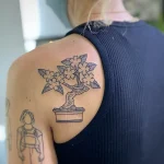 Черно-белая татуировка на лопатке с изображением цветущей сакуры в квадратном горшке, выполненная в стиле минималистичной графики.