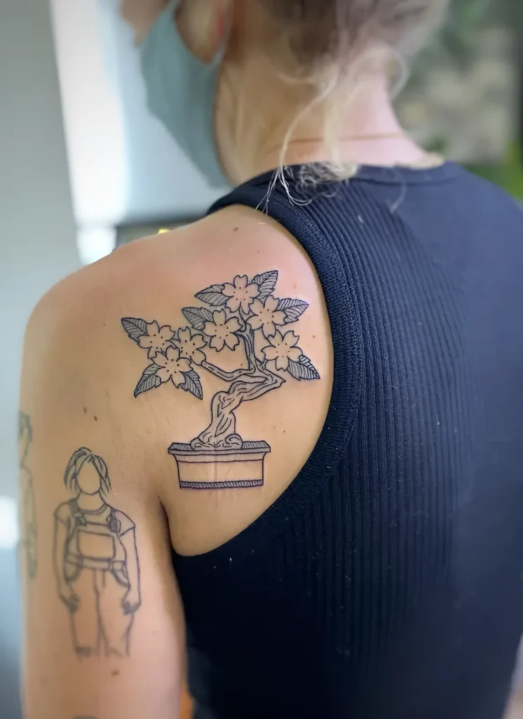 Black and white tattoo on the shoulder blade: a blooming sakura in a square pot, done in a minimalist graphic style.