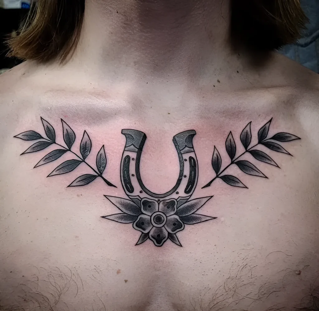 Black and white horseshoe tattoo with flowers on a man's collarbone, done in old-school style with detailed shading.