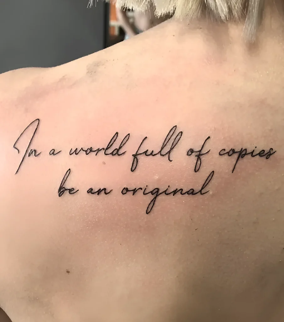 Photo of a tattoo on a man's shoulder blade: black lettering in calligraphic font 'In a world full of copies be an original'.