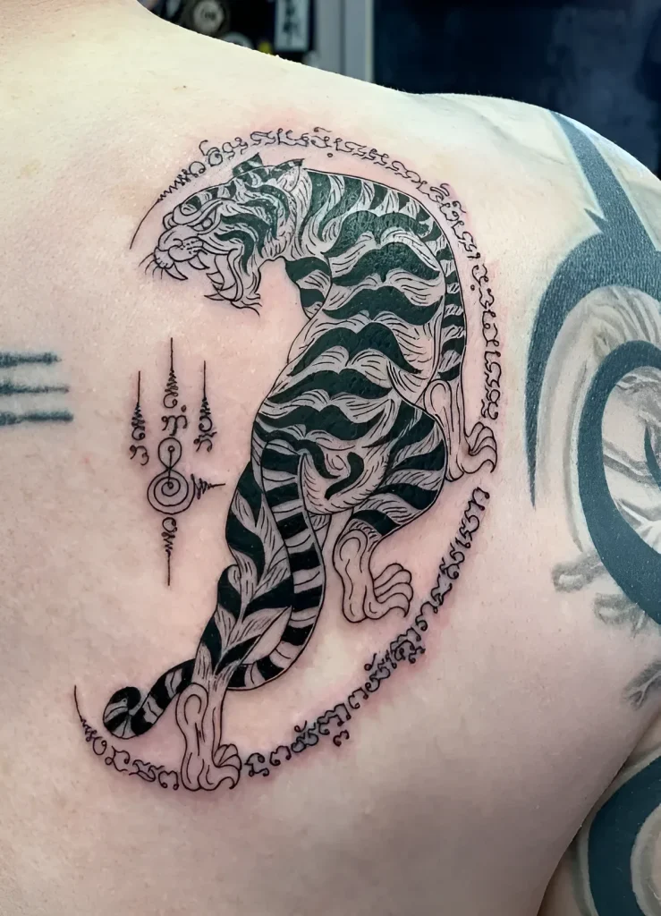 Photo of a tiger tattoo in the Sak Yant style on a man's shoulder blade, done in black ink with Thai symbols.