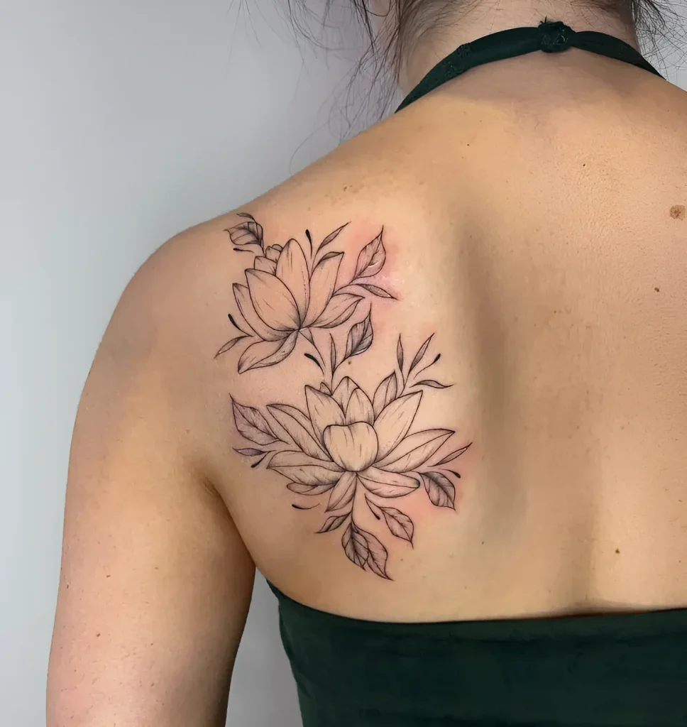 Black and white tattoo of two blooming lotuses with leaves on a woman's shoulder blade, done in a graphic style.