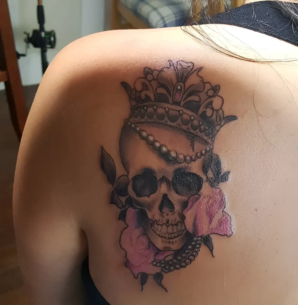 Photo of a tattoo on the shoulder blade: a realistic skull in a crown, adorned with pink roses and a pearl string.