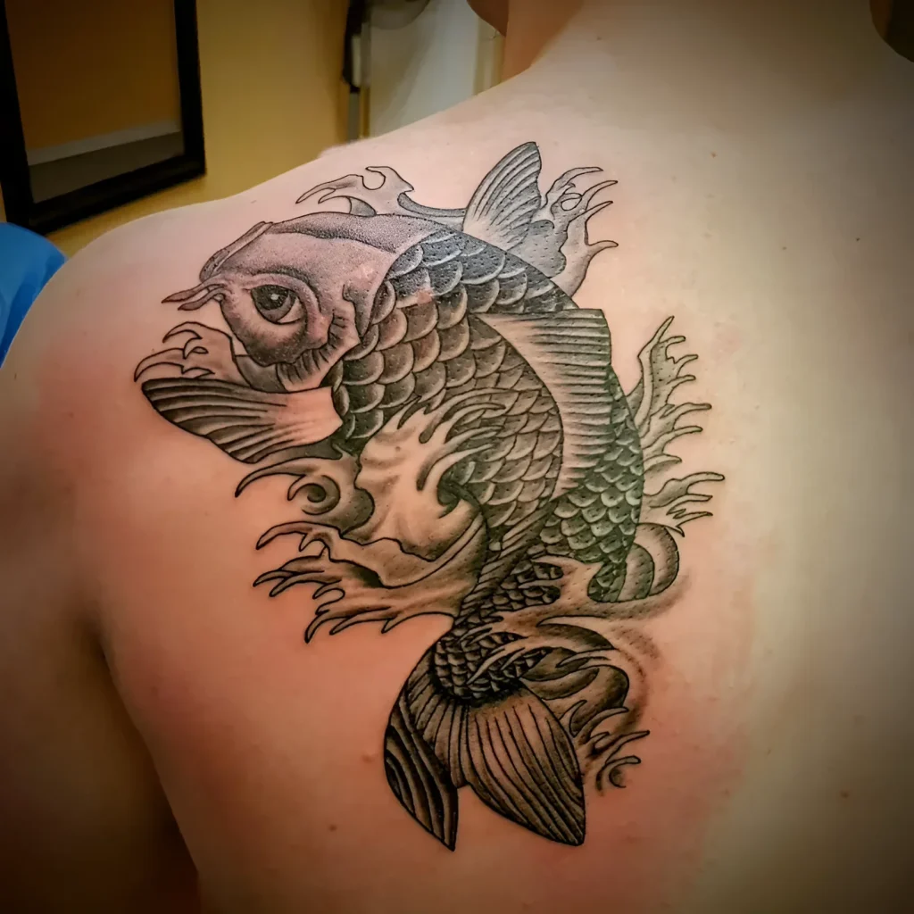 Black and white koi tattoo on the shoulder blade, done in a graphic style with detailed scales and fins.