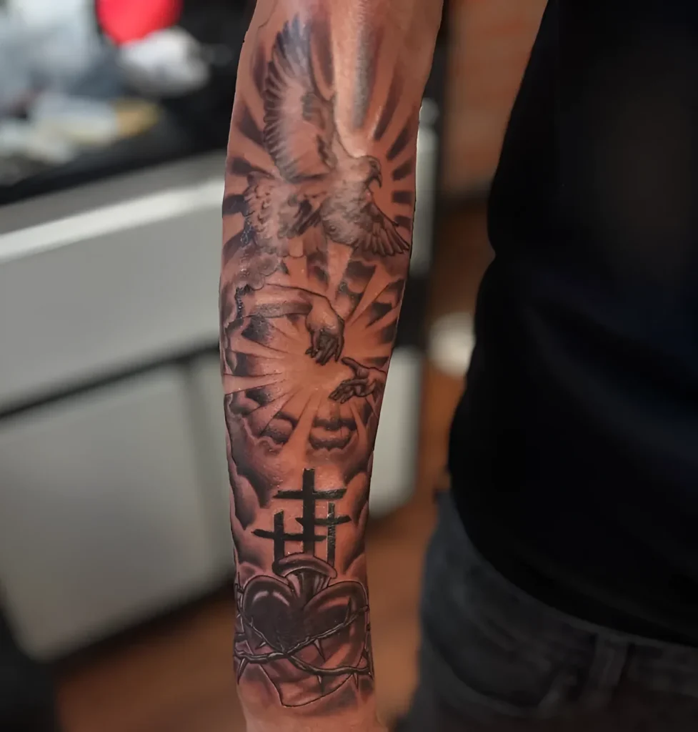 Black and grey forearm tattoo depicting an angel releasing a dove from its hands, and a cross with a crown of thorns, symbolizing spirituality and redemption.