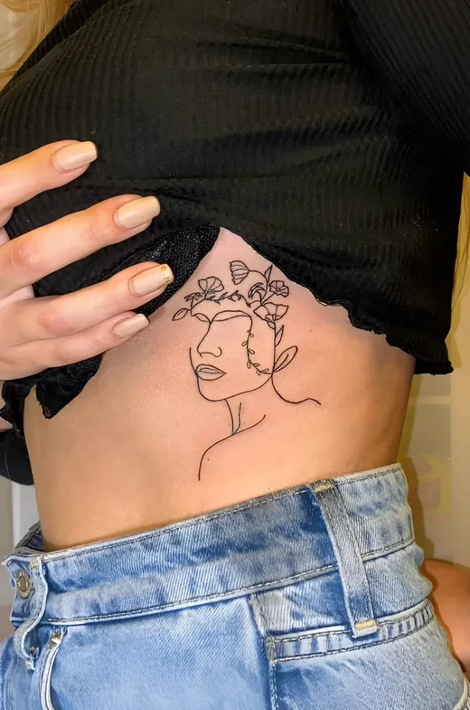 Tatouage sur le flanc d'une jeune femme, réalisé en ligne fine : portrait d'un visage féminin orné de fleurs et de feuilles, dans un style minimaliste.