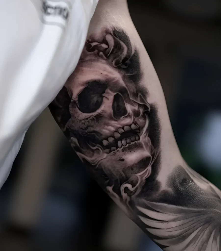 Tatouage noir et blanc d'un crâne sur le biceps, réalisé en technique réaliste avec des effets de fumée et des éléments végétaux ajoutés.