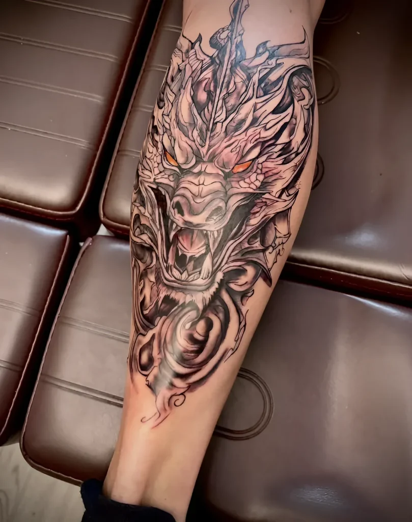 Black and grey dragon tattoo on the calf, detailed face with bared teeth and horns, stylized waves and swirls around.