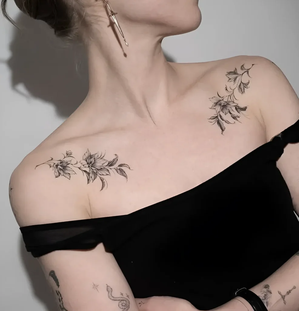 Black and white tattoo of branches with flowers on a girl's collarbone, done in a fine graphic style.