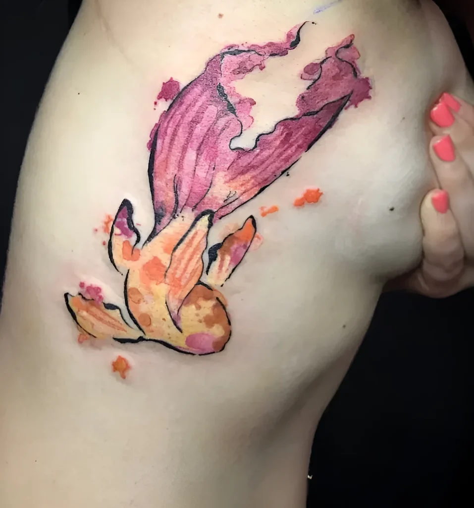 Photo d'un tatouage aquarelle de poisson rouge sur le flanc d'une jeune femme, réalisé dans des tons doux de rose et d'orange.