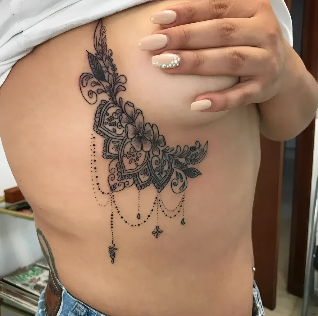 Photo of a tattoo under the female breast: detailed mehndi style ornament with an image of a blooming flower, done in fine line.