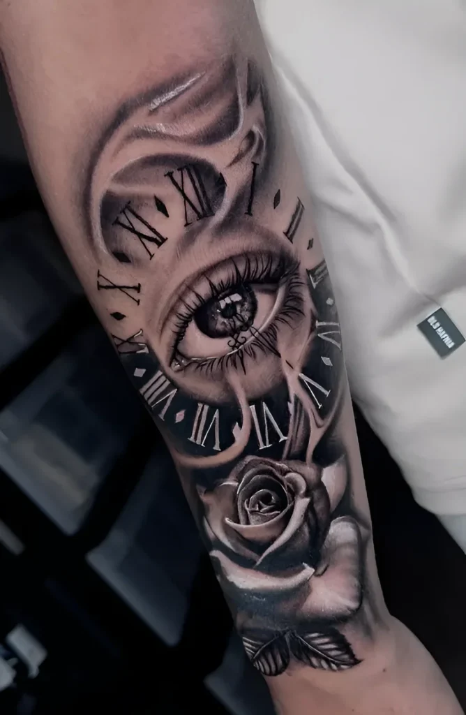 Black and white forearm tattoo: realistic eye enclosed in a clock face with Roman numerals, transitioning into a blooming rose.