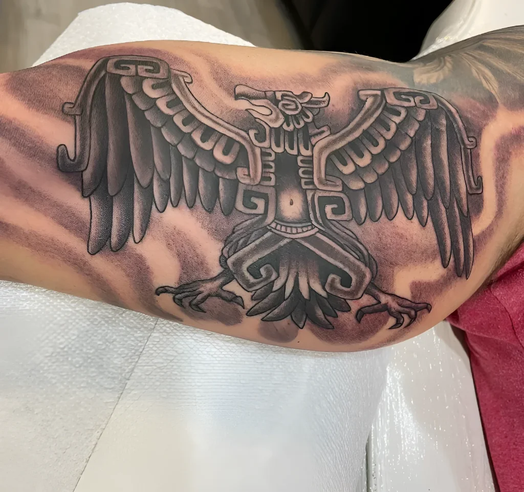 Tatouage noir et gris d'un aigle sur le biceps, réalisé dans un style inspiré de l'art aztèque et maya, avec des plumes détaillées et des motifs géométriques.