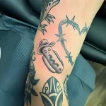 Черно-белая татуировка хот-дога в ковбойской шляпе на руке, выполненная в стиле олд скул с элементами традиционной татуировки.
