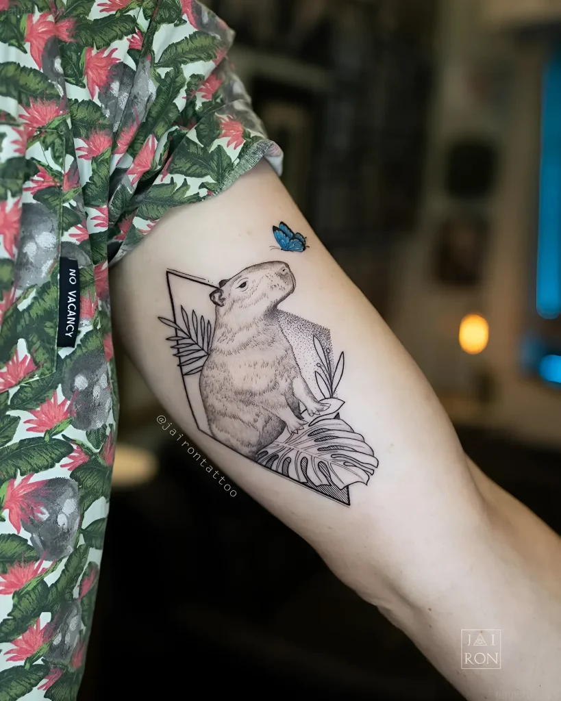 Tatouage noir et blanc d'un capybara dans un cadre géométrique avec un papillon sur l'épaule, réalisé en pointillé.