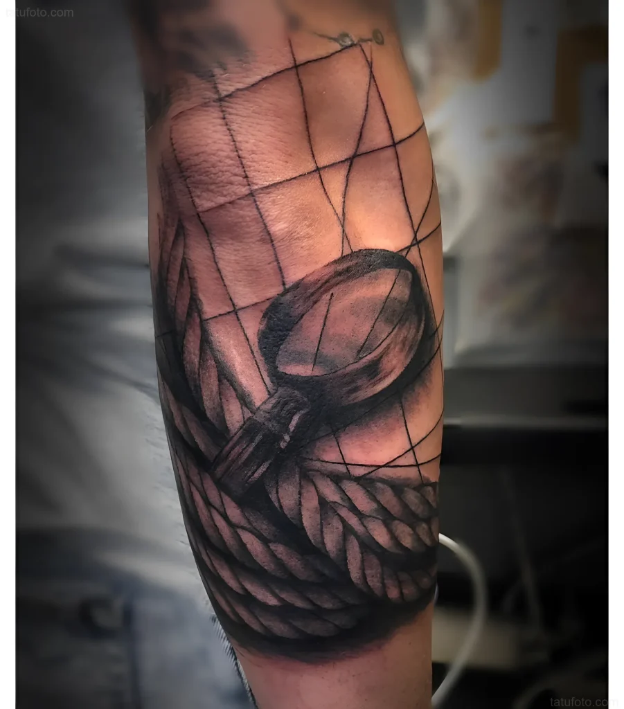Black and grey realistic tattoo of a magnifying glass on an arm entwined with rope, with a geometric grid background.