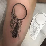 Черно-белая татуировка лупы, обвитой нежными цветами, на внутренней стороне плеча.