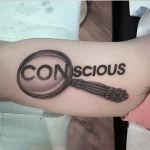 Черно-серая татуировка лупы, увеличивающей слово 'Conscious' на руке, символизирующая осознанность и внимательность.