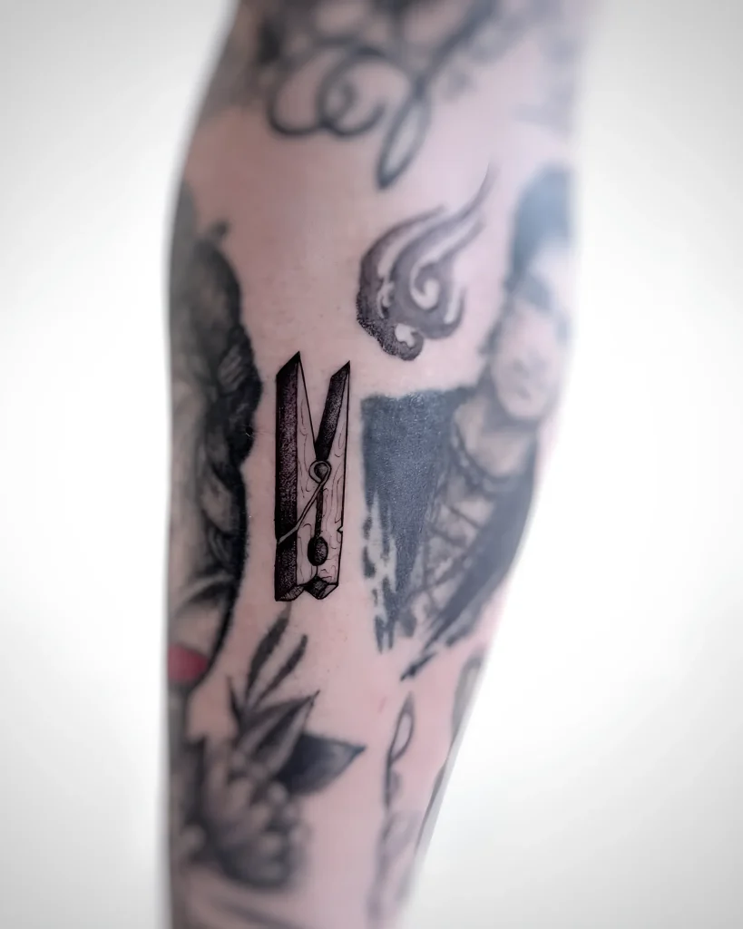 Tatouage en noir et blanc d'une pince à linge sur le bras, réalisé dans un style graphique avec un travail détaillé sur la texture du bois et du métal.