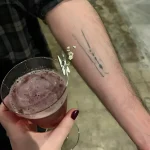 Минималистичная татуировка прищепки на предплечье, выполненная в технике лайнарт, рядом с бокалом коктейля.
