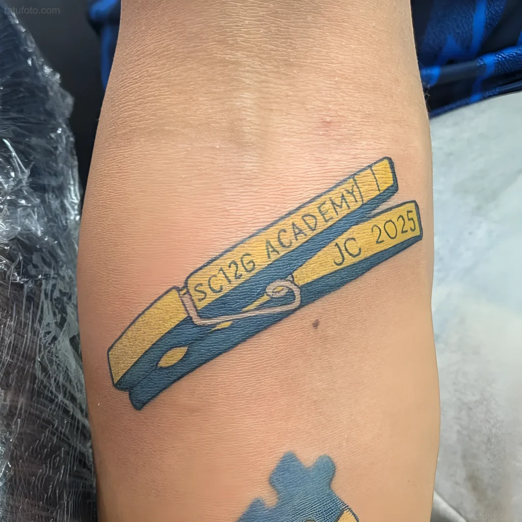 Tatouage d'une pince à linge avec l'inscription 'SC12G ACADEMY JC 2025' sur le bras, réalisé dans un style minimaliste.