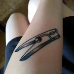 Черно-белая татуировка прищепки на внутренней стороне бедра, выполненная в минималистичном стиле.