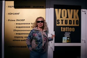 Женщина в солнцезащитных очках стоит перед входом в тату-студию "Vovk Studio", на стене слева указаны услуги студии.