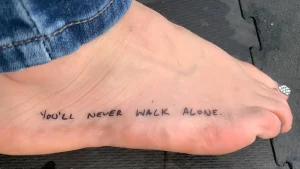 Крупный план стопы с татуировкой фразы 'You'll Never Walk Alone' черными чернилами.
