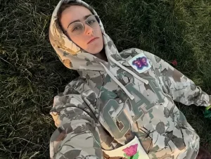 Элла Эмхофф в камуфляжном худи GAP лежит на траве, смотрит прямо в камеру.