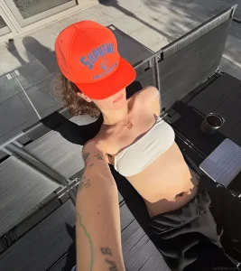 Элла Эмхофф в оранжевой кепке Supreme и белом топе-бандо загорает на шезлонге, демонстрируя татуировки на руке.