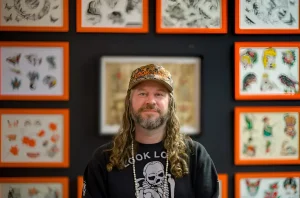 CJ Fishburn, тату-мастер и владелец Look Look Tattoo, в Солт-Лейк-Сити, стоит перед стеной, украшенной винтажными эскизами татуировок в оранжевых рамках.