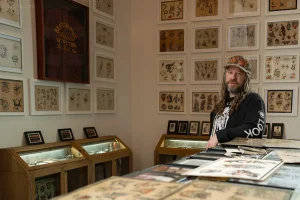 Татуировщик CJ Fishburn стоит в своем магазине и музее Look Look Tattoo в Солт-Лейк-Сити, окруженный стенами, украшенными старинными эскизами татуировок.