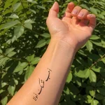 Фотография татуировки на запястье: арабская каллиграфия с надписью «أحببتك أولاً» на нежной женской руке, окруженной зеленью.