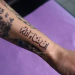 Чёрная татуировка надписи 'إن شاء الله' (Инша Аллах) на предплечье, выполненная тонкой линией.