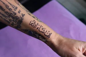 Чёрная татуировка надписи 'إن شاء الله' (Инша Аллах) на предплечье, выполненная тонкой линией.