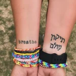 Фото татуировки: Надпись 'breathe.' на запястье и ивритская фраза 'Тиккун Олам' на внутренней стороне запястья, выполненные черными чернилами.