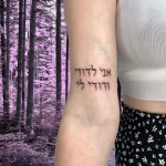 Фотография татуировки на внутренней стороне предплечья: черная надпись на иврите «אני לדודי ודודי לי» на фоне размытого леса.